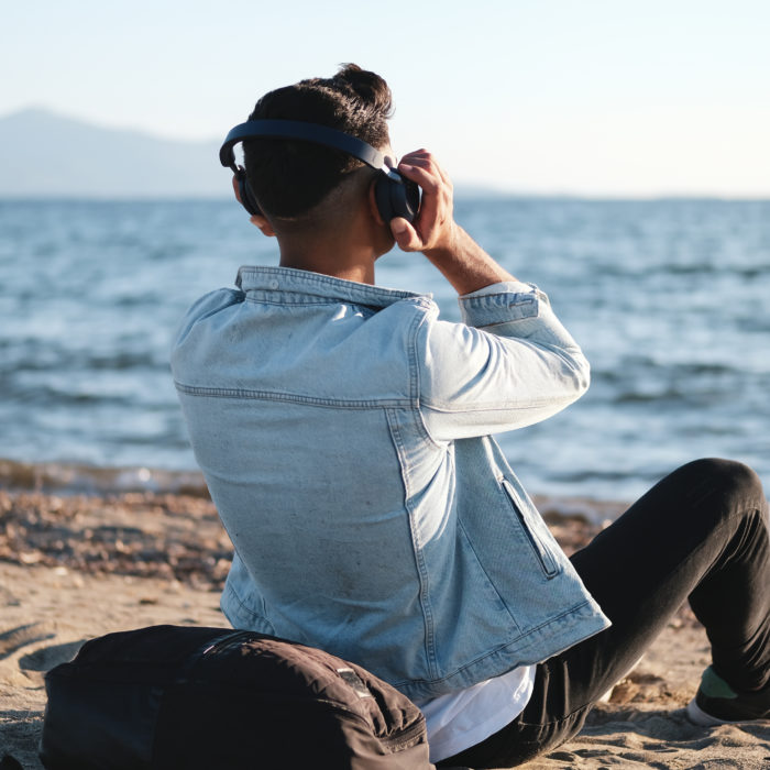 Young man sitting at beach with headphones, listening music. He is listening to a podcast and watching the ocean in front of him